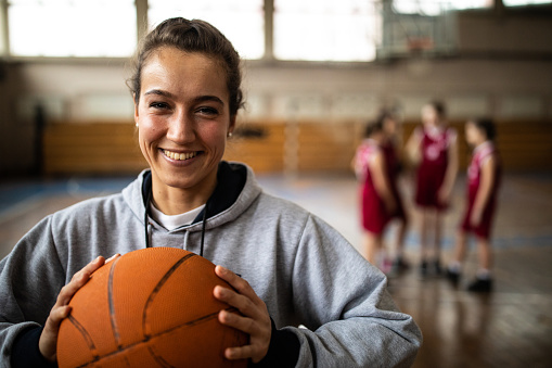 Young beautiful female basketball coach standing in school gym , holding basketball ball , looking at camera and smiling