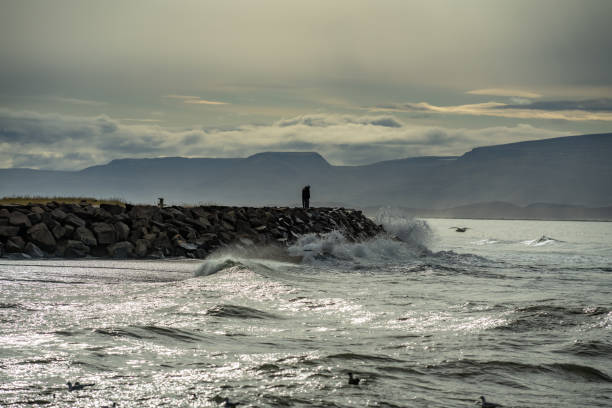Man sea storm beach. The element large waves break on the stone shore. Seascape with an impending storm. A man poses against the background of the raging surf. Resort concept, climate weather changes