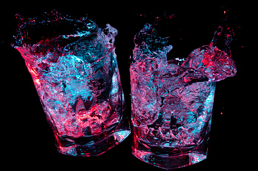 Two rocks glasses clinking in toast while glowing colorful liquid splashes out on a black background