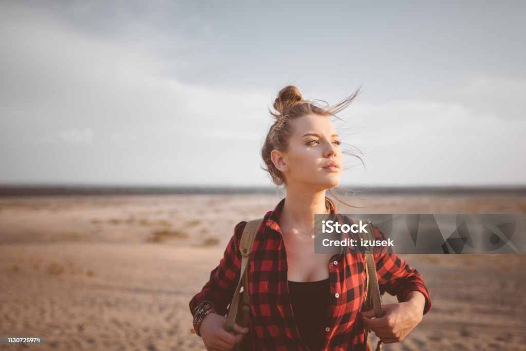 Young woman looking away in desert against sky Thoughtful blond young woman standing against sky. Beautiful hipster is hiking in desert. Female explorer is looking away. Hair Bun Stock Photo