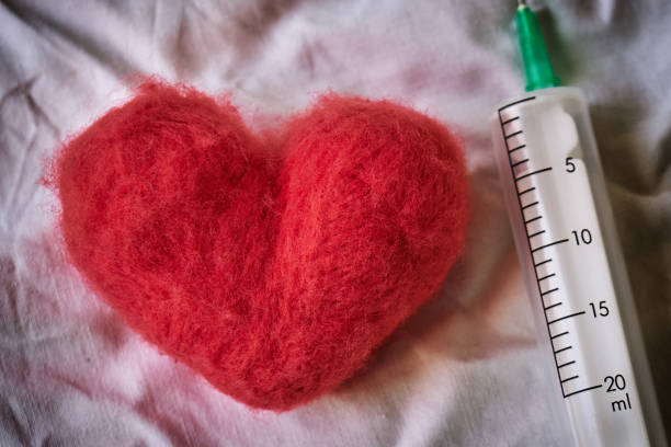 Red wool felted heart and a syringe lying on a white crumpled bed sheet. Top view. Heart treatment concept