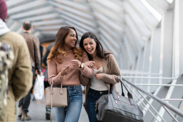 Two (2) female friends wait for their plane Two (2) young female travelers laugh as they wait for their flight to arrive. They are fashionable and enjoying their time together. airports canada stock pictures, royalty-free photos & images