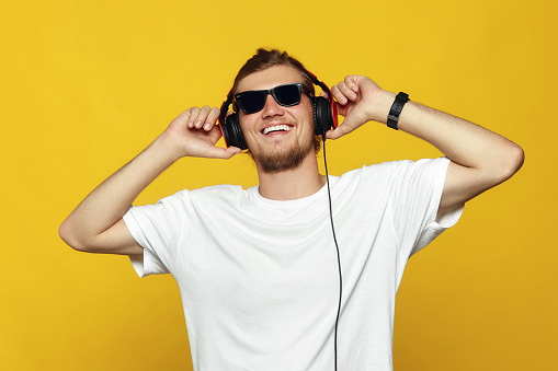 Close up portrait of a cheerful young caucasian male with sunglasses listening to music with headphones isolated over yellow background. Handsome hipster in white t-shirt listening music.