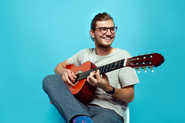 guapo joven hipster con anteojos tocando una guitarra mientras se sienta aislado sobre fondo azul. - plucking an instrument fotografías e imágenes de stock