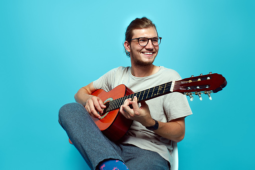 Portrait of young guy with eyeglasses playing a guitar while sitting isolated over blue background.
