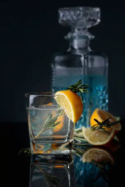Cocktail gin-tonic with lemon slices and twigs of rosemary on a black reflective background.