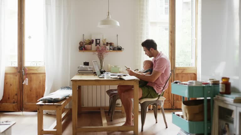 Side view of man sitting with son at table in kitchen. Boy is playing with toy on father's lap. They are spending leisure time at home.