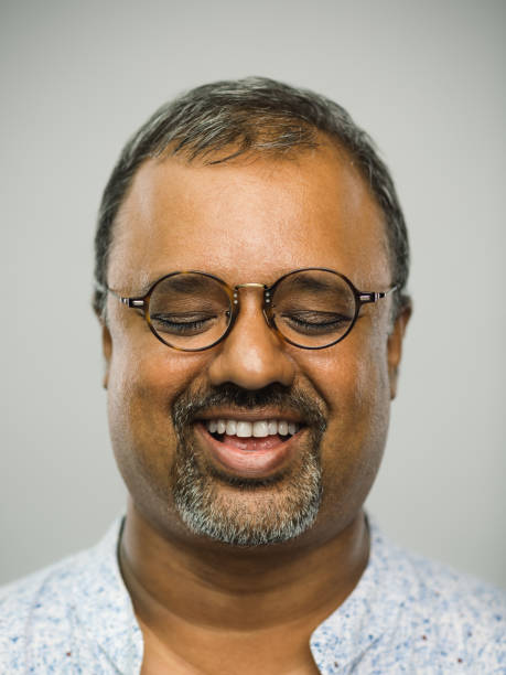 Real indian mature man with excited expression and eyes closed Close up portrait of indian mature man with excited expression and eyes closed against white gray background. Vertical shot of real people resting and laughing in studio with glasses and beard. Photography from a DSLR camera. Sharp focus on eyes. chubby arab stock pictures, royalty-free photos & images