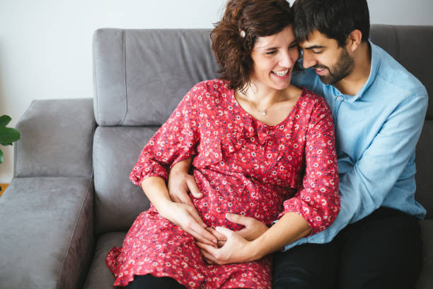 Loving happy pregnant couple at home Happy pregnant couple embracing lovingly on the couch at home 8 months pregnant stock pictures, royalty-free photos & images
