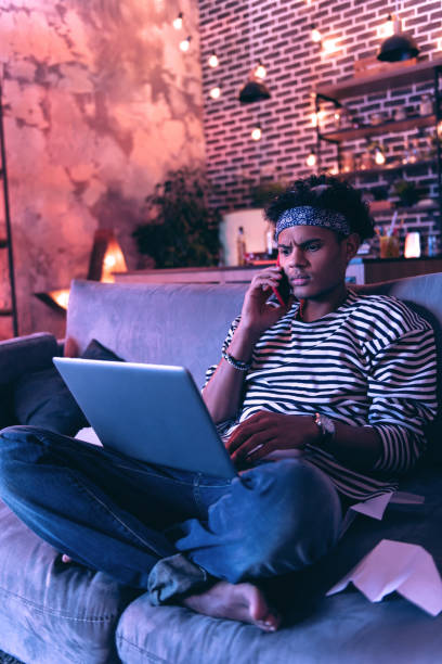 istock African American boy sitting barefoot on the couch and checking his mail while talking on the phone. 1130760553