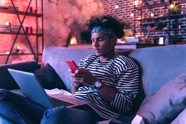 istock Young man sitting on sofa holding a laptop on legs and typing a message to girlfriend. 1130760554