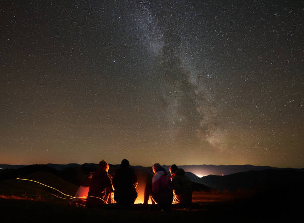 Friends resting beside camp, campfire under night starry sky Back view group of four young friends hikers resting together around bonfire near camp and tourist tent at night in the mountains. On background beautiful night starry sky full of stars and Milky way. campfire stock pictures, royalty-free photos & images