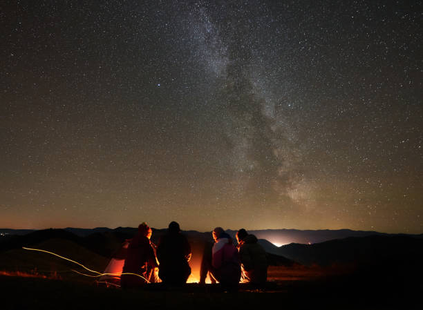 Friends resting beside camp, campfire under night starry sky Back view group of four young friends hikers resting together around bonfire near camp and tourist tent at night in the mountains. On background beautiful night starry sky full of stars and Milky way. camp fire stock pictures, royalty-free photos & images