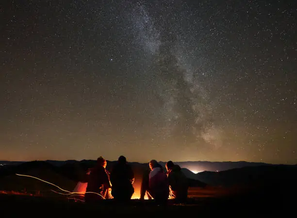 Back view group of four young friends hikers resting together around bonfire near camp and tourist tent at night in the mountains. On background beautiful night starry sky full of stars and Milky way.