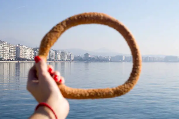 womans hand with red maniqure holding the greek bagel koulouri with sea and city on the background.