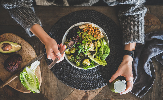Healthy dinner, lunch setting. Flat-lay of vegan championship game or Buddha bowl with hummus, vegetable, salad, beans, couscous and avocado, green smoothie and woman's hands over wooden table, top view