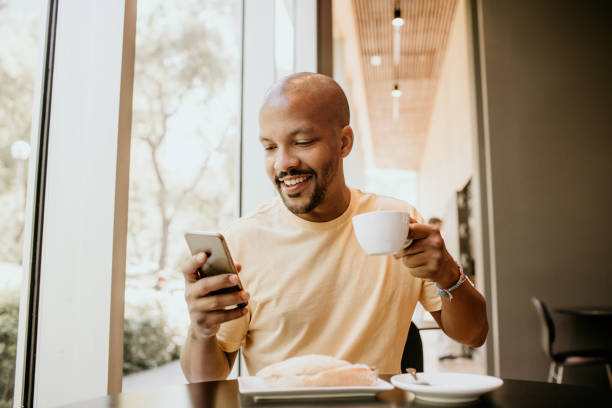 istock Happy cheerful African hipster holding mug, drinking fresh cappuccino, browsing internet and checking newsfeed on social media.Man using cell phone during coffee break at modern cafe. 1130796457