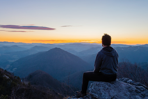 Hiker sitting alone on top of the hill waiting for sunrise and enjoying scenic view of hills and valleys below. Hiking, achievement, expectation, optimism and self-reflection concepts.
