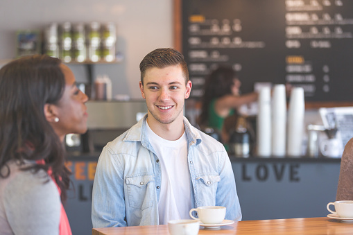 Two friends, a Caucasian male and African American female in their 20s, drink coffee and hang out together in a modern coffee shop. The focus is on him as he listens to her.