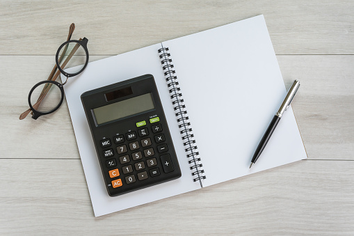 White opening blank paper note book with pen, calculator and eyeglasses on light grey wooden table background with copy space using as finance, debt and budget plan.