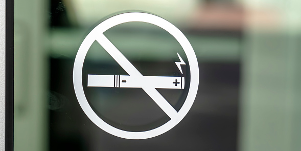 No Smoking sign symbol on a glass surface. Close up view of a No Smoking sign on a transparent glass surface of a building. The symbol specifically prohibits the use of electronic cigarettes.