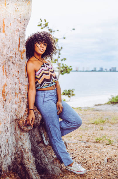 Girl Power Beautiful african american woman with big natural hair leans against a tree wearing a crop top, jeans and sneakers. She exudes an aura of power, fierceness, and confidence. Her outfit is retro and she could be from any decade. She is clearly a force crop top view stock pictures, royalty-free photos & images
