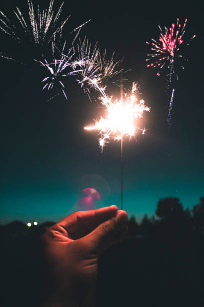 Hand Holding Sparkler with Fireworks in the Background A hand holding a sparkler with fireworks going off in the background at night victoria day canada photos stock pictures, royalty-free photos & images