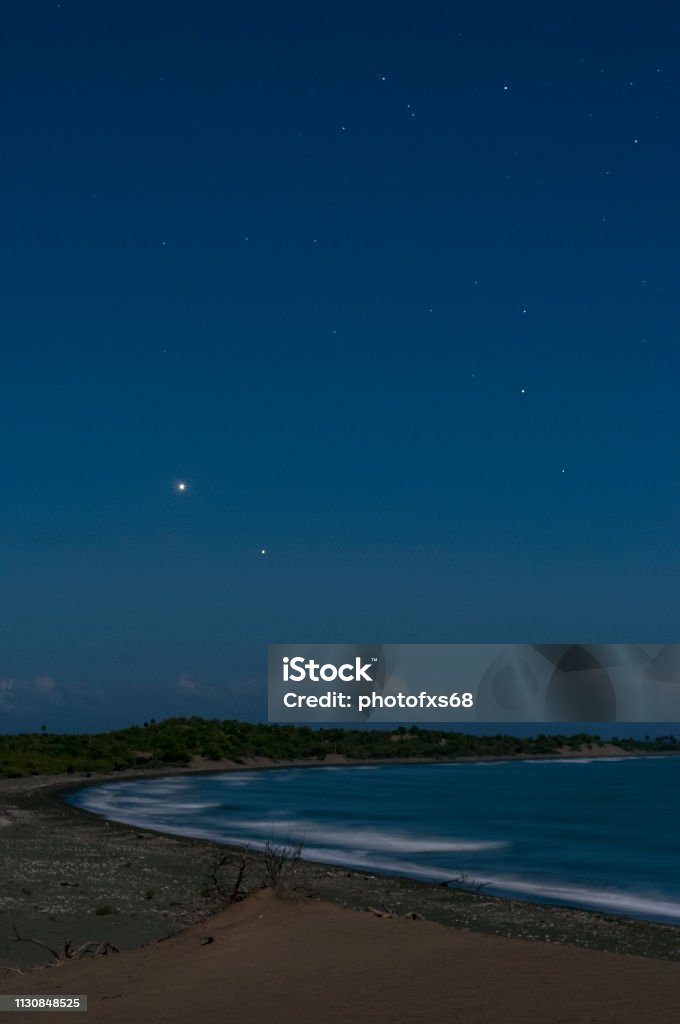 Venus and Jupiter conjunction over the beach Scene of Venus and Jupiter in conjunction coming out of the east, just above the dunes of Bani in the Dominican Republic, the scene is described during the early morning just before dawn, you can see the sea and the waves breaking on the shore where you can see many stones Some clouds are seen on the horizon Arid Climate Stock Photo