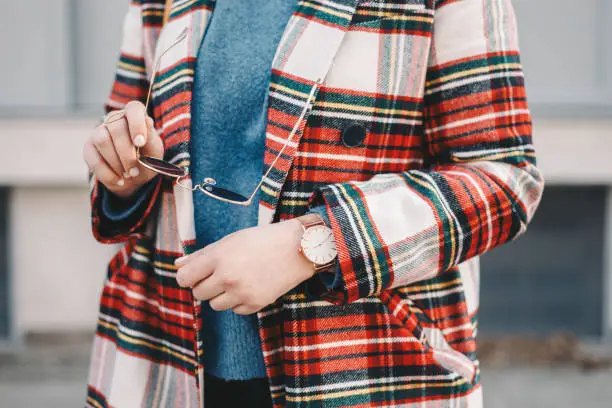 Photo of Close-up detail of fashion accessories, stylish young woman wearing an overcoat with a tartan pattern, a golden ring, a pair of modern sunglasses and a wrist watch.