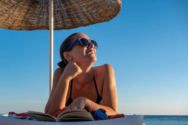 schöne frau liest buch auf dem liegen am strand - sonnenbaden stock-fotos und bilder