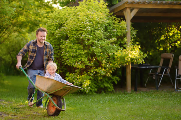 szczęśliwy mały chłopiec bawiący się w taczki pchającej tatę w ogrodzie domowym w ciepły słoneczny dzień. - farmer farm family son zdjęcia i obrazy z banku zdjęć