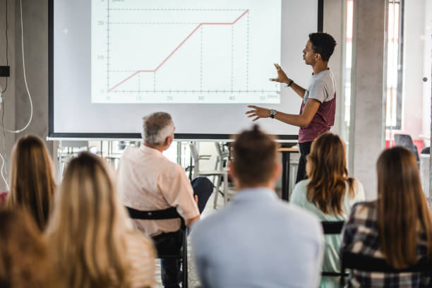 Black university student giving a presentation during an education event. African American college student looking at projection screen while holding a presentation in lecture hall. lecture hall training classroom presentation stock pictures, royalty-free photos & images