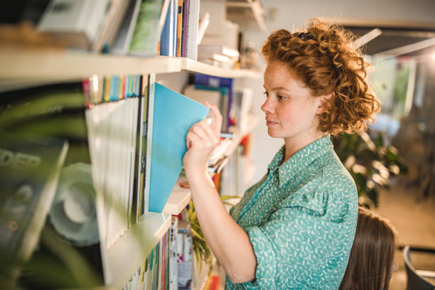 istock Redhead woman taking book from the shelf in a library. 1130890991