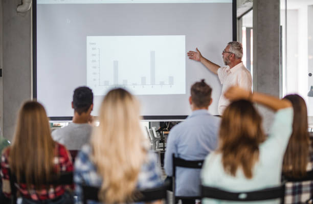 Senior teacher giving a presentation to group of university students at lecture hall. Mature professor teaching group of college students while aiming at projection screen at lecture hall. teacher projector stock pictures, royalty-free photos & images