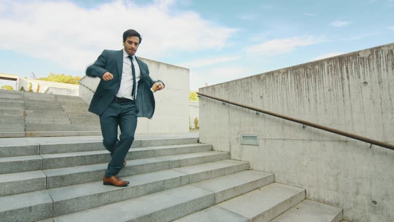 Cheerful and Happy Businessman in a Suit is Holding Coffee and Actively Dancing While Walking Down the Stairs. Scene Shot in an Urban Concrete Park Next to Business Center. Sunny.