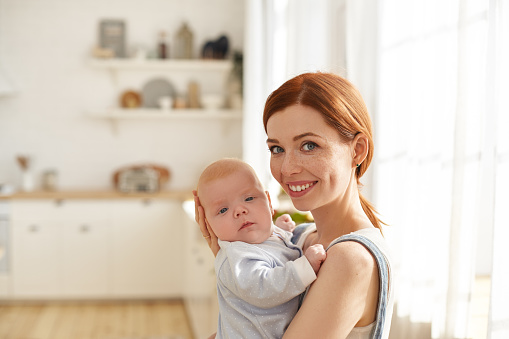 Indoor image of attractive charming young mom with ginger hair and freckles nursing her cute baby, singing him to sleep, walking around in spacious living room with infant in her arms and smiling