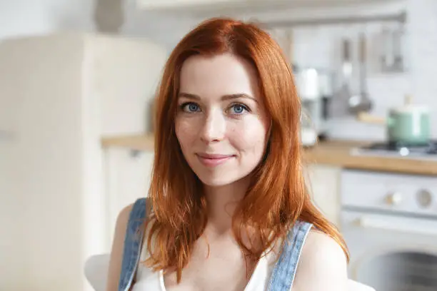 Photo of Headshot of charming beautiful young European woman with red shiny hair and freckles cooking or having meal in kitchen interior, looking at camera with cute smile. People and lifestyle concept
