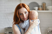 Image of happy blue eyed young mother with ginger hair looking and smiling joyfully at camera, embracing her unrecognizable baby in blue bodysuit. Childcare, motherhood and family bonding concept