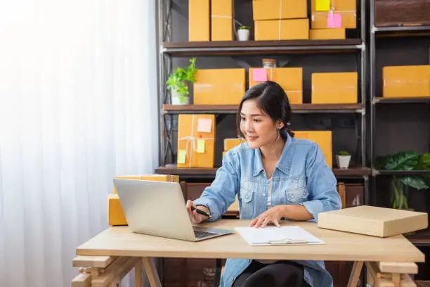 Photo of Young Asian female freelancer using her laptop and working at home, Small business female owner or Start up small business entrepreneur working online marketing packaging box delivery