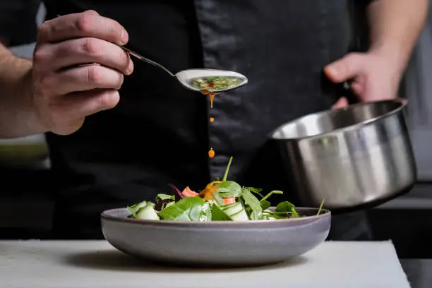 Photo of Close-up of the hands of a male chef on a black background. Pour sauce from the spoon on the salad dish.