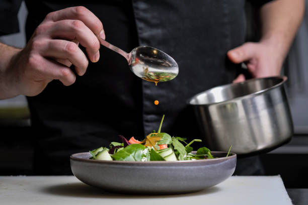 primo piano delle mani di uno chef maschio su sfondo nero. versare la salsa dal cucchiaio sul piatto di insalata. - chef foto e immagini stock