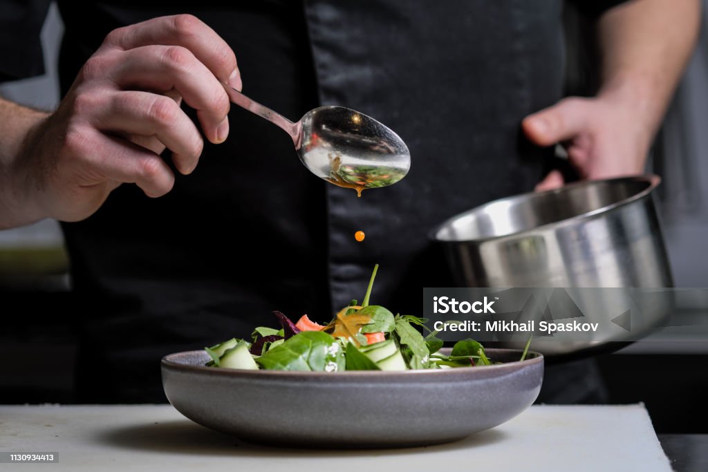 Close-up of the hands of a male chef on a black background. Pour sauce from the spoon on the salad dish. Close-up of the hands of a male chef on a black background. Pour sauce from the spoon on the salad dish. Food decoration. Chef Stock Photo