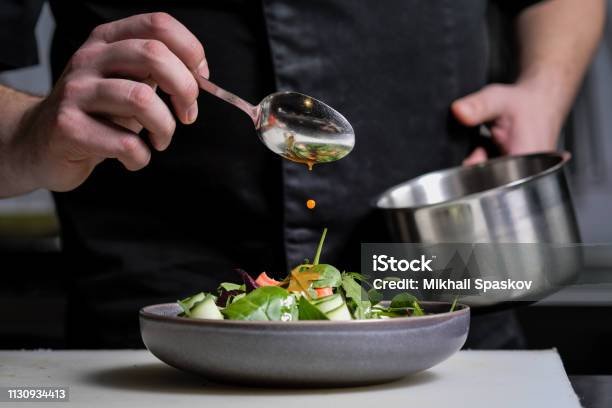 Closeup Of The Hands Of A Male Chef On A Black Background Pour Sauce From The Spoon On The Salad Dish Stock Photo - Download Image Now