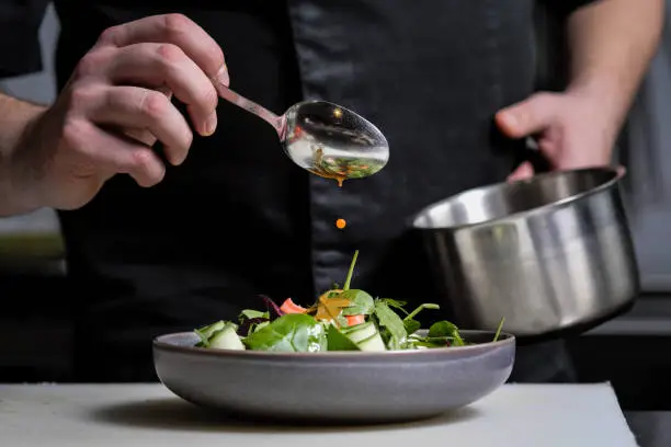 Photo of Close-up of the hands of a male chef on a black background. Pour sauce from the spoon on the salad dish.