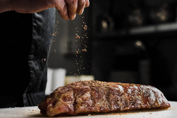 Raw piece of meat, beef ribs. The hand of a male chef puts salt and spices on a dark background. stock photo
