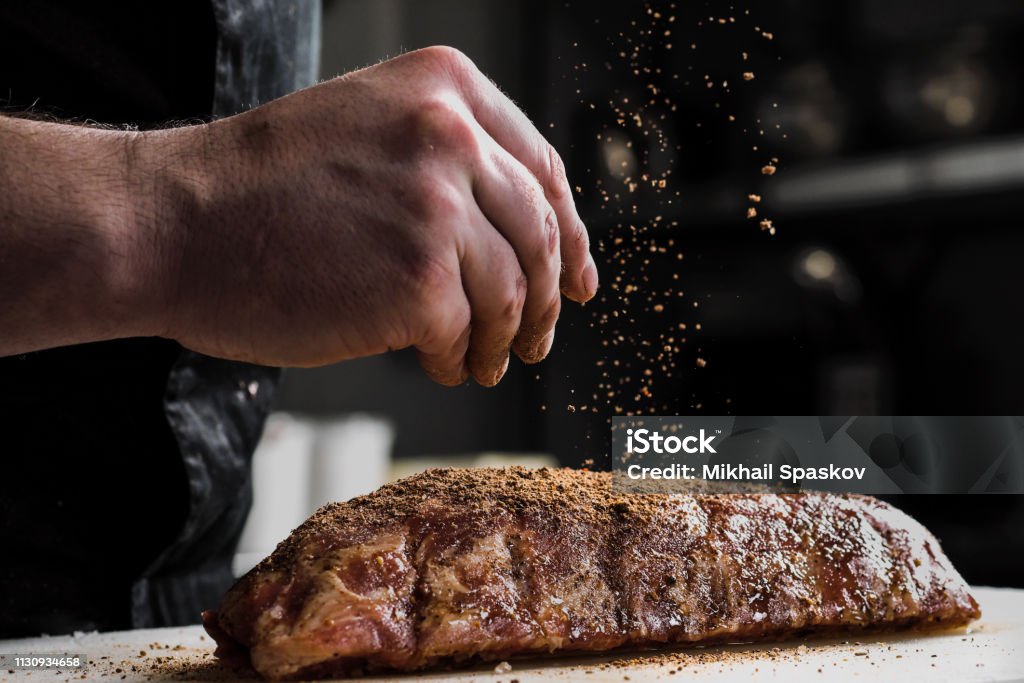 Trozo de carne cruda, costillas de ternera. La mano de un cocinero macho pone sal y especias sobre un fondo oscuro. - Foto de stock de Carne libre de derechos