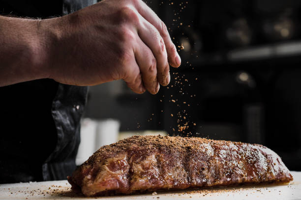 trozo de carne cruda, costillas de ternera. la mano de un cocinero macho pone sal y especias sobre un fondo oscuro. - sal condimento fotografías e imágenes de stock