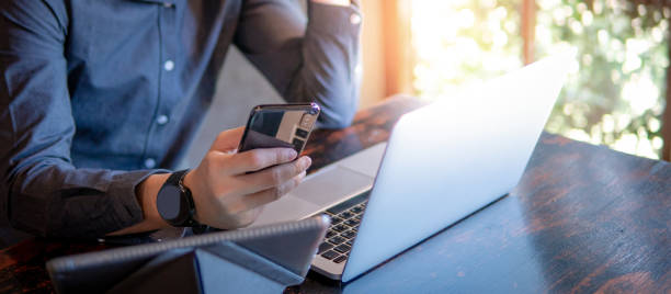 スマートフォンを保持する男性の手。カフェで仕事をしながら、ラップトップコンピュータとデジタルタブレットを使用してビジネスマン。モバイルアプリやモノのインターネットの概念。� - 保護 写真 ストックフォトと画像