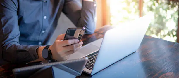 Photo of Male hand holding smartphone. Businessman using laptop computer and digital tablet while working in the cafe. Mobile app or internet of things concepts. Modern lifestyle in digital age.