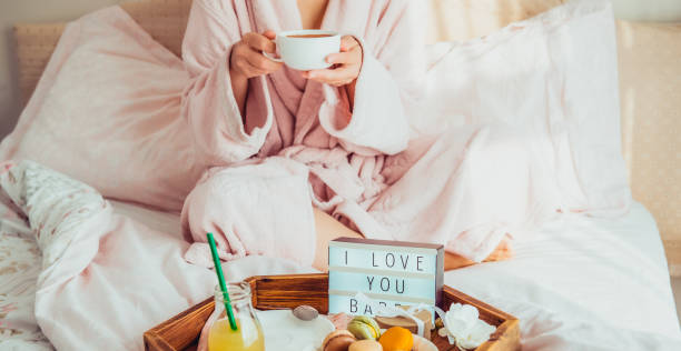 istock Romantic Breakfast in bed with I love you text on lighted box, macaroons, gift box on tray and blurred cropped woman in a bathrobe drinking her coffee. Birthday, Valentine's day morning. Wide banner. 1130942945